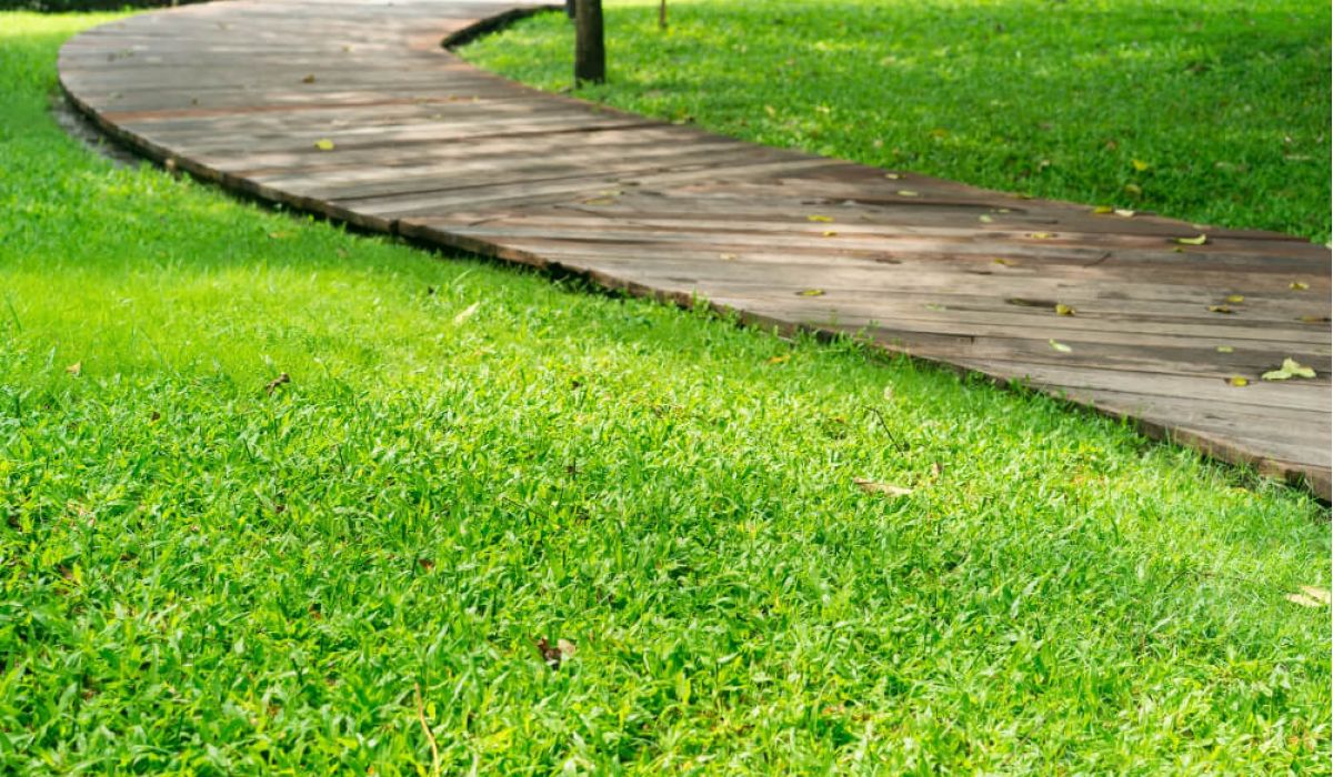A gently winding wooden boardwalk curves through a well-maintained green lawn in a park-like setting. The path is bordered by lush grass, with a few trees and colorful flower beds visible in the background.