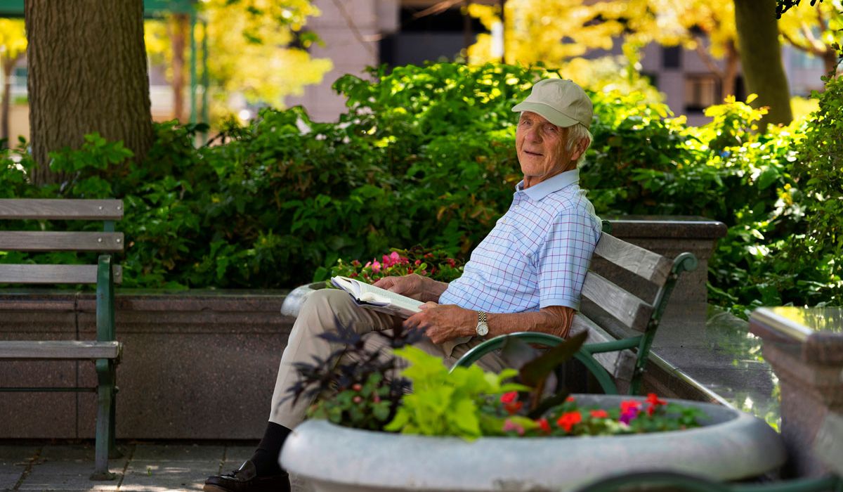 An older man wearing a light-colored cap and a short-sleeved checkered shirt sits on a park bench reading a book.