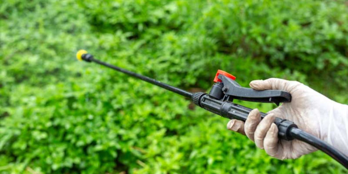 A gloved hand holds a spray lance with a trigger handle, aimed toward dense green vegetation. The image shows the application of a treatment or spray in an outdoor garden setting.