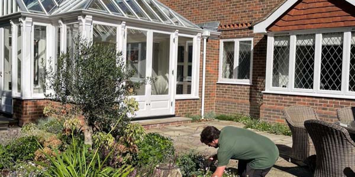 A gardener kneels beside a flowering garden bed in a paved courtyard, tending plants outside a brick house with a white conservatory.