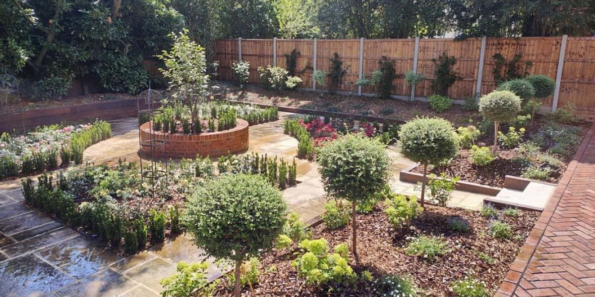 Structured ornamental garden with geometric planting beds arranged around a circular brick planter. Clipped topiary trees, flowering borders, stone paving, and timber fencing create a formal, symmetrical design.