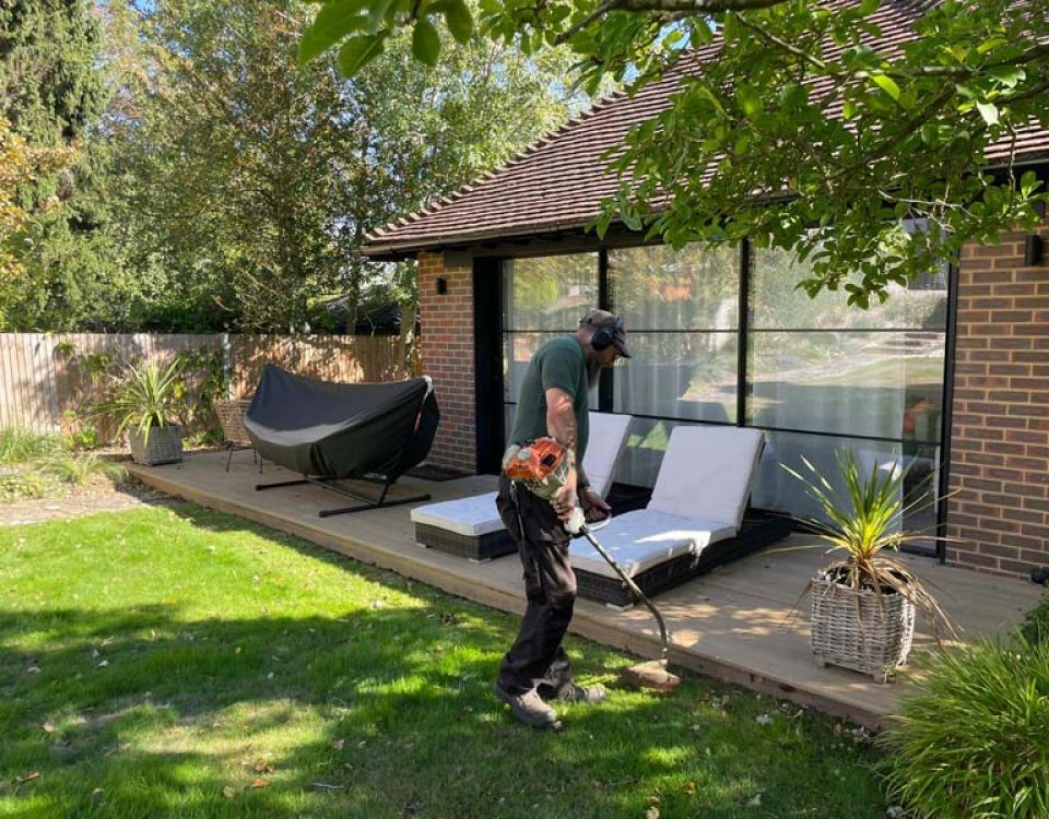 A grounds maintenance worker uses a grass trimmer along the edge of a lawn beside a modern brick building. Outdoor furniture sits on a patio behind them, with trees providing partial shade.
