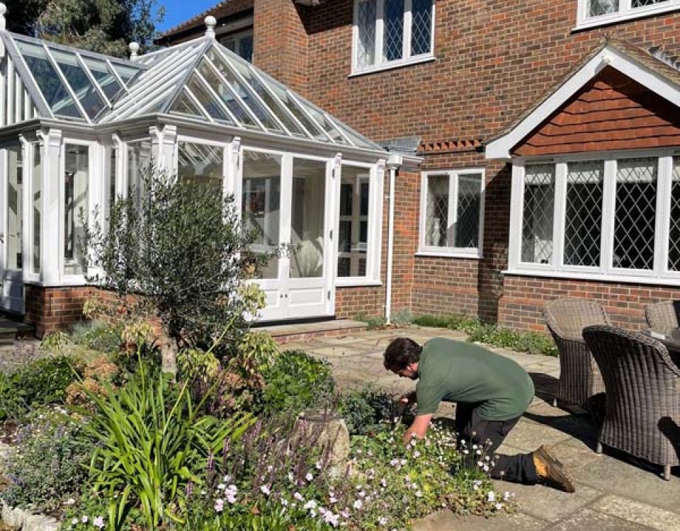 A gardener kneels beside a flowering garden bed in a paved courtyard, tending plants outside a brick house with a white conservatory.