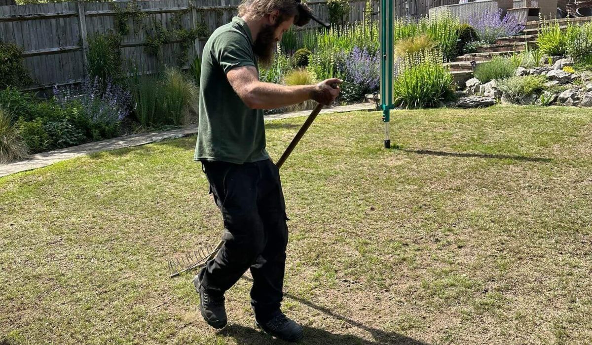 A person in a green polo shirt and dark work trousers rakes a dry, patchy lawn in a sunny landscaped garden.