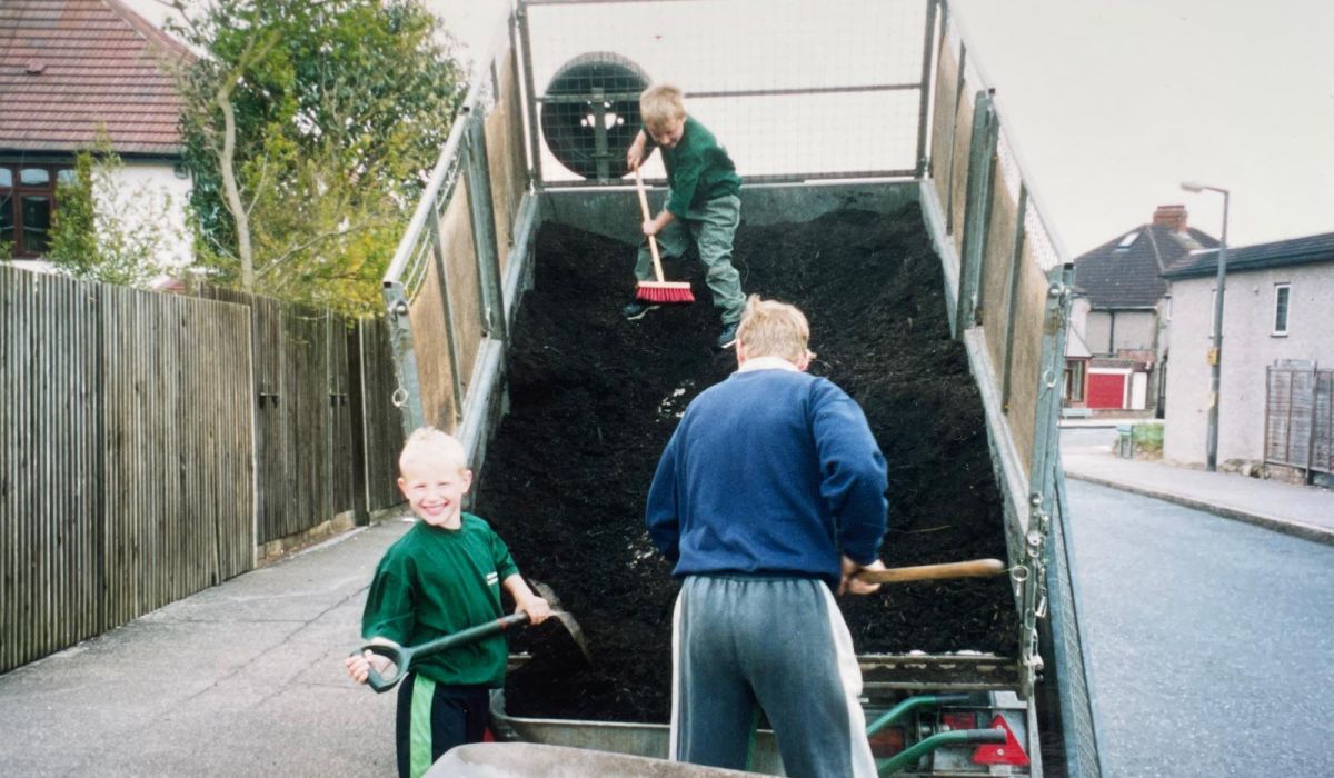 Adult and two children unloading compost from the back of a tipper truck using shovels and a wheelbarrow.