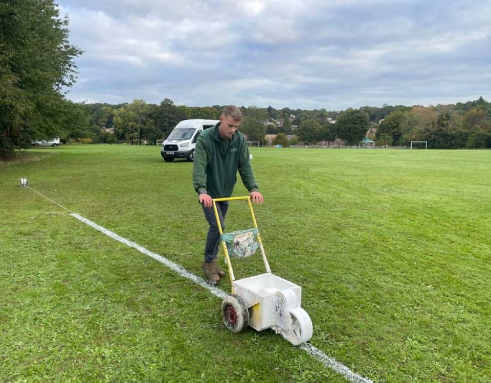 A groundskeeper pushes a wheeled line-marking machine across a sports field, painting a white boundary line on the grass. A white van, goalposts, and trees are visible in the background beneath a cloudy sky.