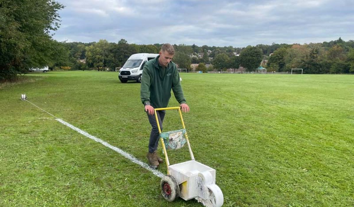 A groundskeeper pushes a wheeled line-marking machine across a sports field, painting a white boundary line on the grass. A white van, goalposts, and trees are visible in the background beneath a cloudy sky.