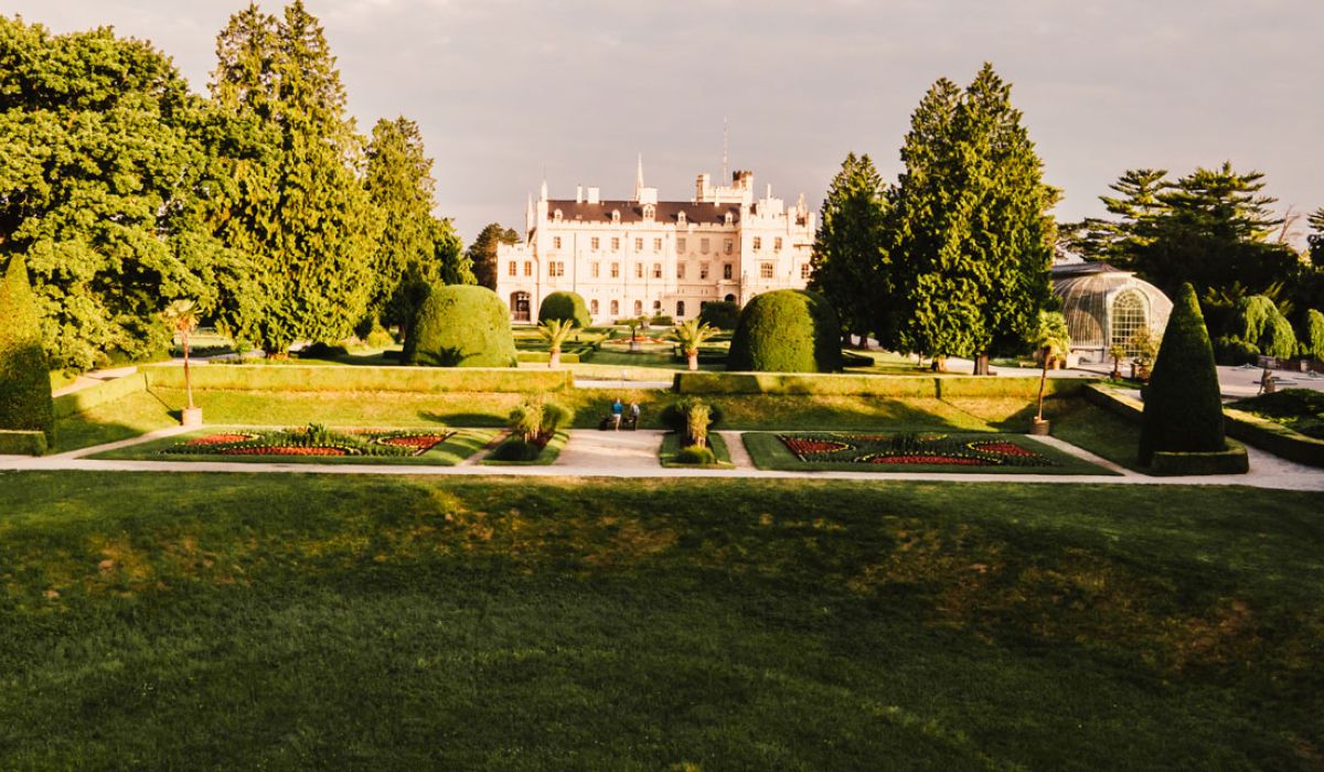 A grand estate with a large historic mansion sits beyond a formal landscaped garden. Symmetrical lawns, trimmed hedges, flower beds, and manicured topiary line the grounds, while tall mature trees frame the scene.