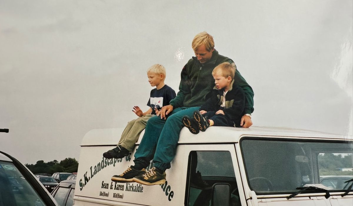 Adult and two young boys sitting on top of an S.K. Landscapes & Maintenance service vehicle at an outdoor event.