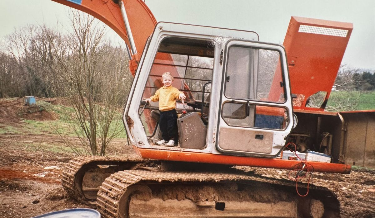 Young child standing in the cab of an excavator on a construction site, smiling during an early introduction to plant machinery.
