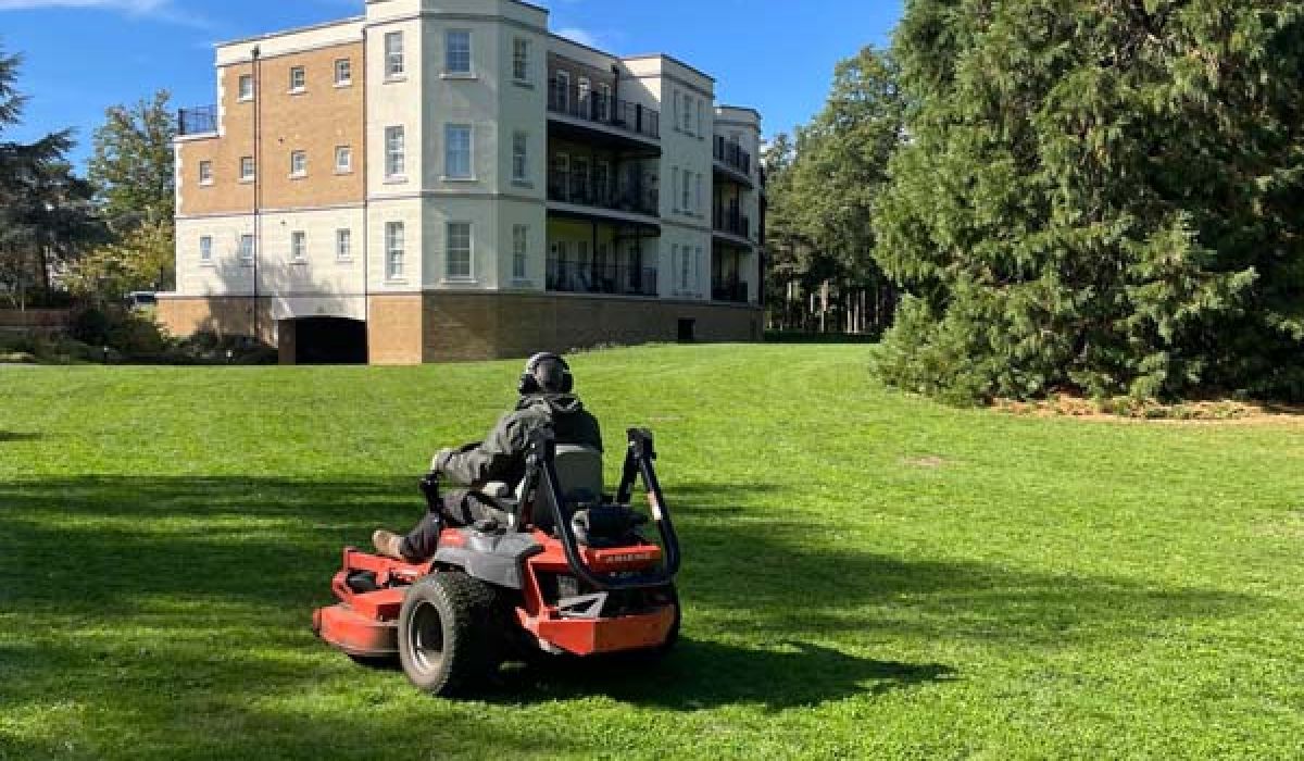 A grounds maintenance worker drives a ride-on lawn mower across a large green lawn in front of a residential apartment building. Mature trees and a bright blue sky frame the scene.