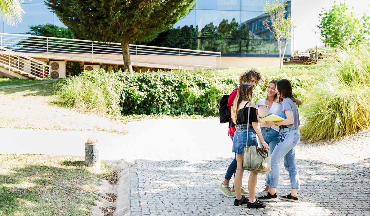 Four students stand together on a paved walkway outside a modern campus building, chatting and smiling while holding notebooks and a tablet.
