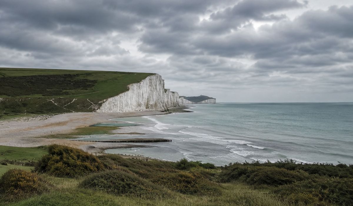 Wide coastal landscape in East Sussex showing the white chalk cliffs of the Seven Sisters stretching along the shoreline beneath a dramatic overcast sky.