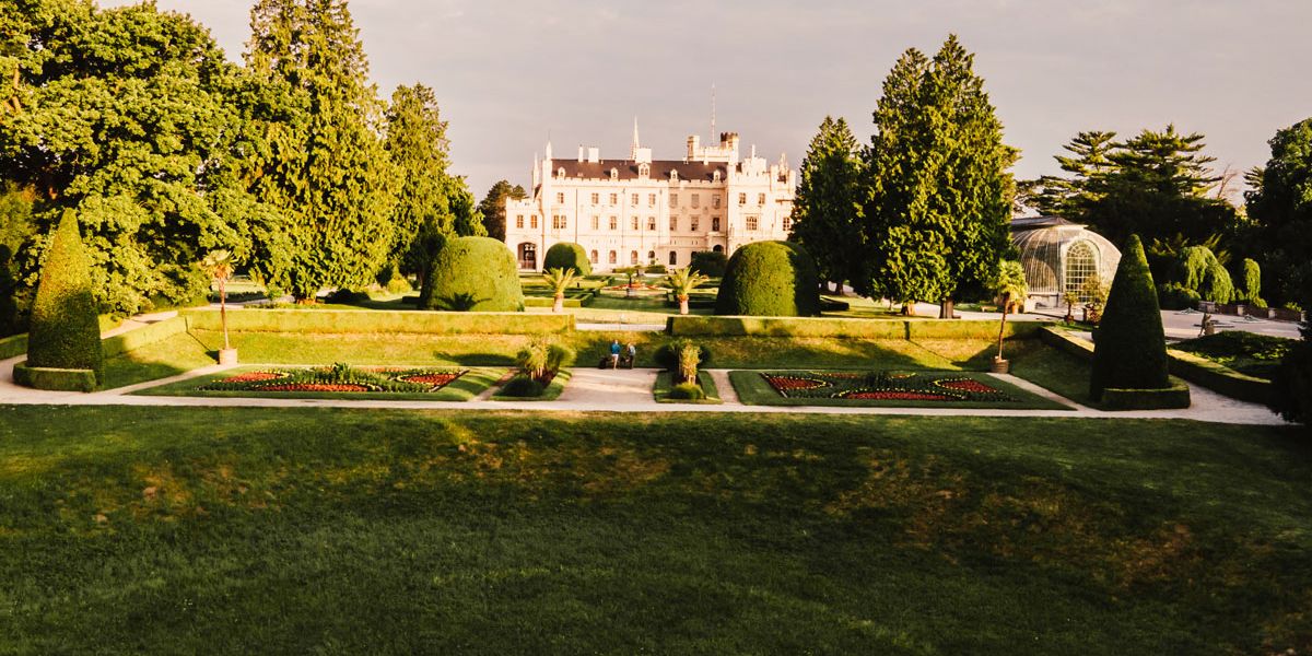 A grand estate with a large historic mansion sits beyond a formal landscaped garden. Symmetrical lawns, trimmed hedges, flower beds, and manicured topiary line the grounds, while tall mature trees frame the scene.