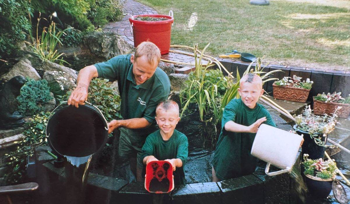 Adult and two children emptying buckets into a garden pond, working together on pond maintenance.
