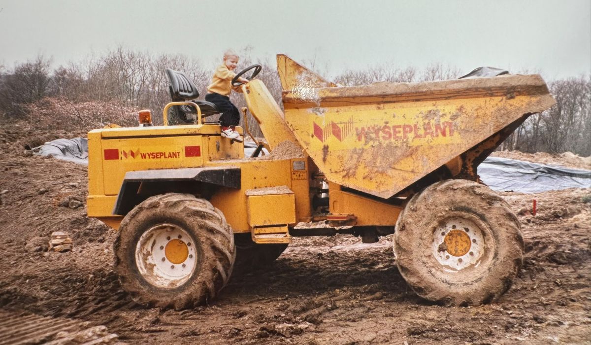 Young child sitting in the driver's seat of a dumper truck on a muddy construction site.