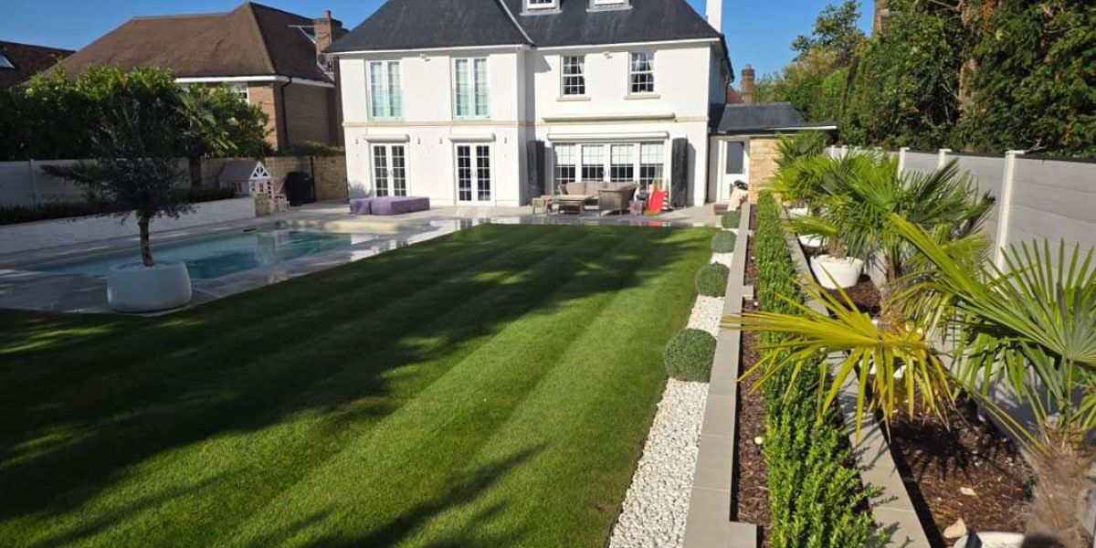 Large luxury garden with a formal striped lawn leading to a swimming pool and elegant white house. Architectural planting beds with palms, clipped shrubs, and decorative stone borders line the lawn.
